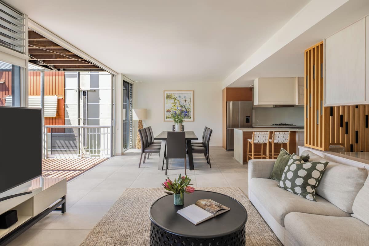 A modern living room with a sofa, coffee table, TV, dining area, kitchen, and a staircase leading to an outdoor balcony.