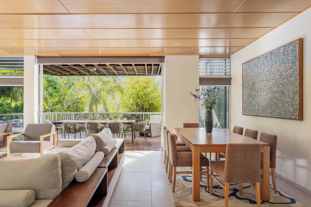 A modern dining area with a wooden table and chairs, a sofa, and a view of greenery through a large window.