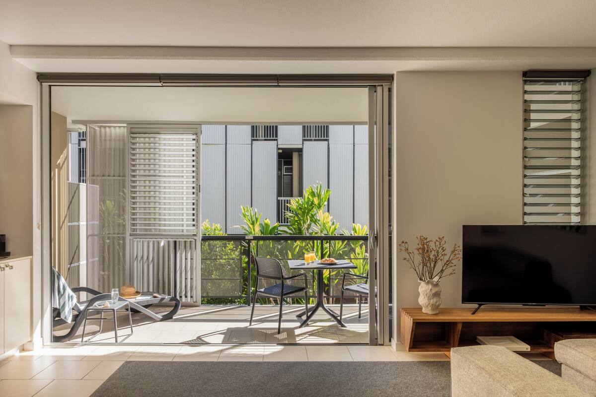 A modern living room with a TV, a sofa, a balcony with a table and chairs, and a sliding glass door leading to a balcony with plants.