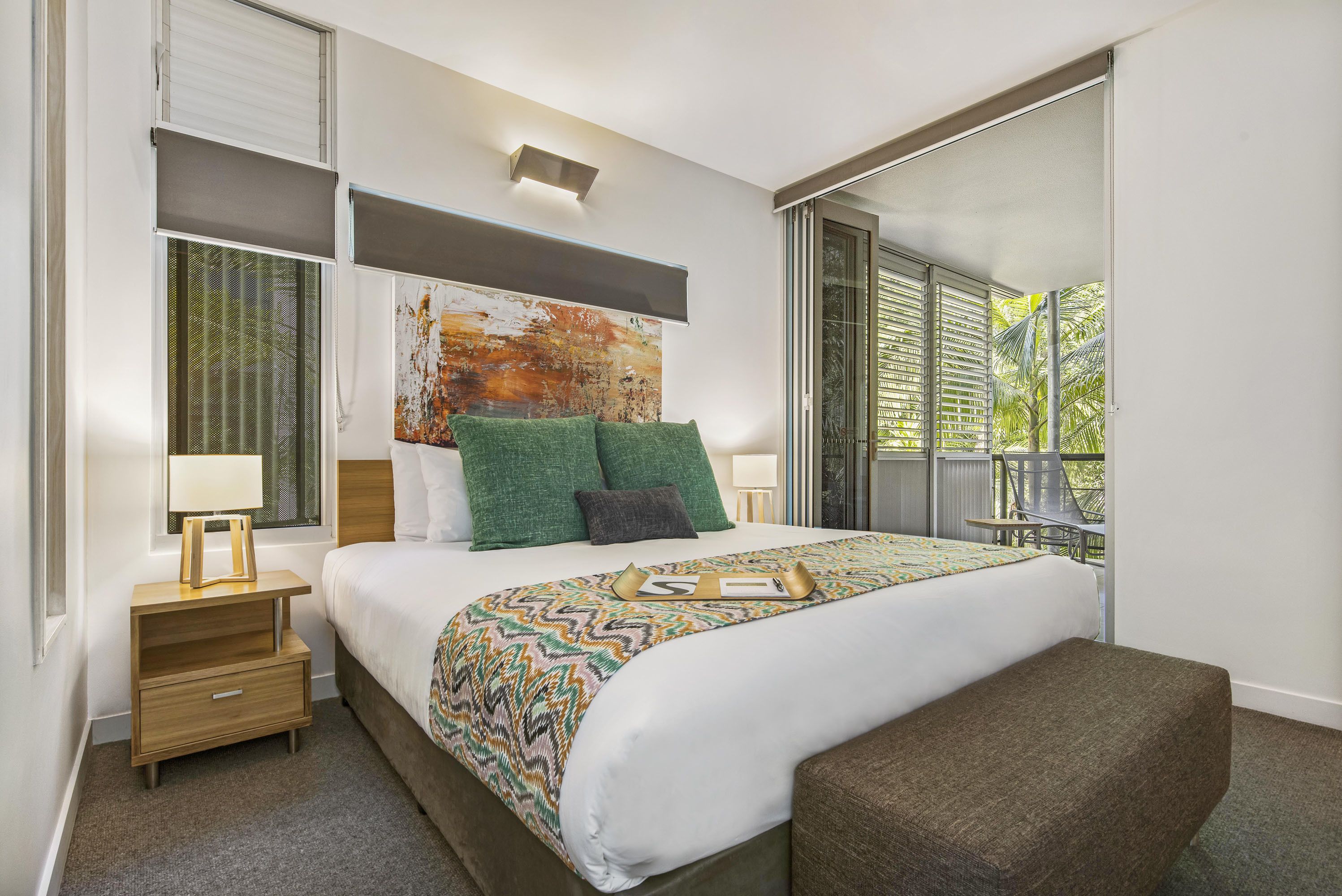 A modern hotel room with a large bed, a nightstand, a bench, and a view of the outdoors.