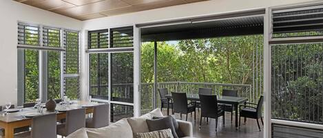 Una sala de estar moderna con sofá, mesa de comedor y sillas, ventanas amplias con vista a zonas verdes y techo de madera.