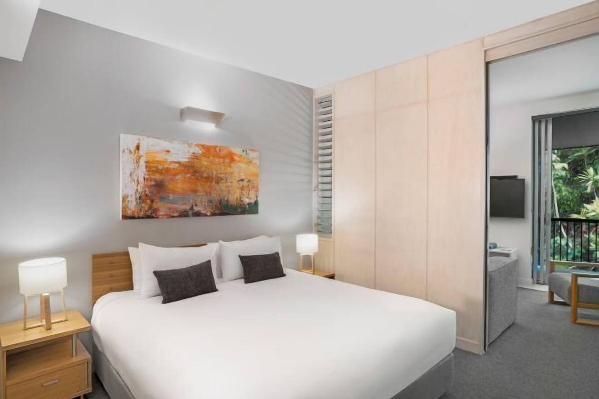 A modern hotel room with a large bed, a painting on the wall, a TV, and a balcony with plants.