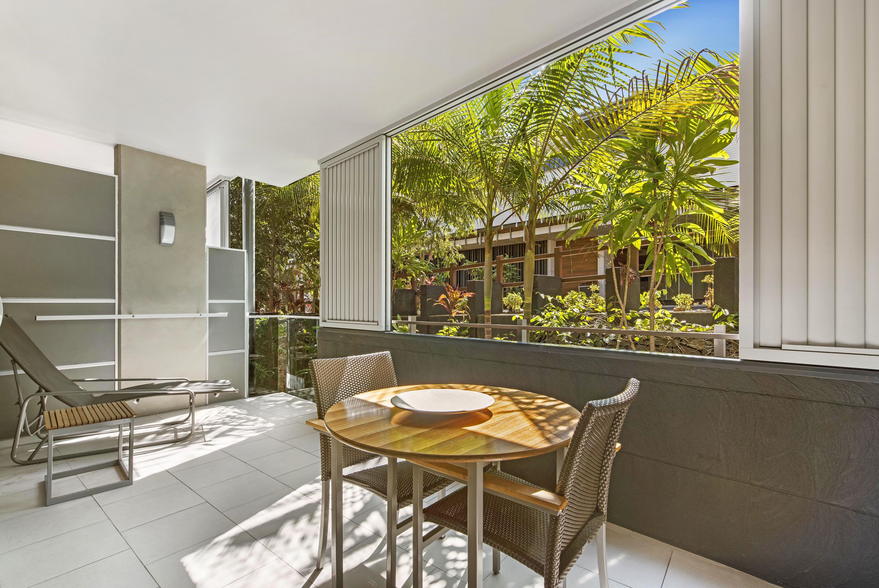 A sunlit balcony with a dining table and chairs, a lounge chair, and a view of a lush garden and buildings.