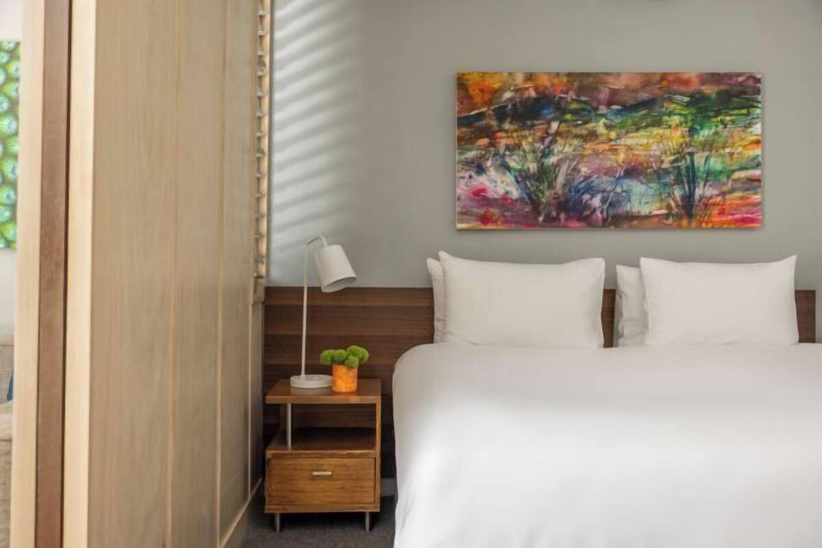 A hotel room with a bed, a nightstand with a lamp and a plant, a colorful abstract painting on the wall, and a wooden wardrobe.