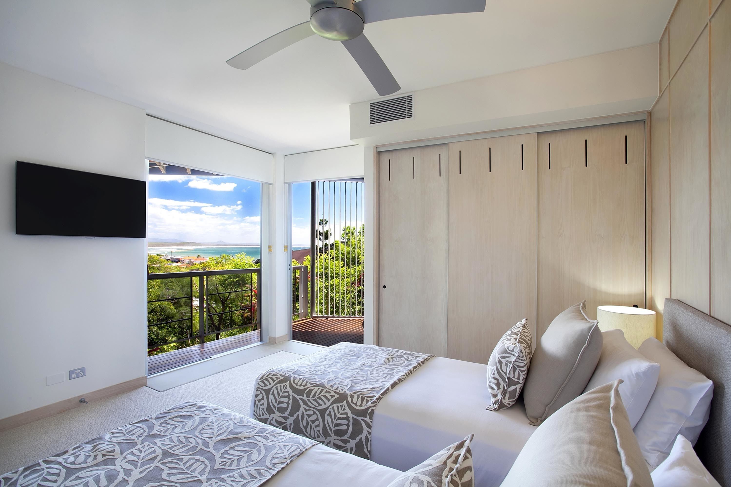 A modern bedroom with a large window, a ceiling fan, and a view of the ocean.