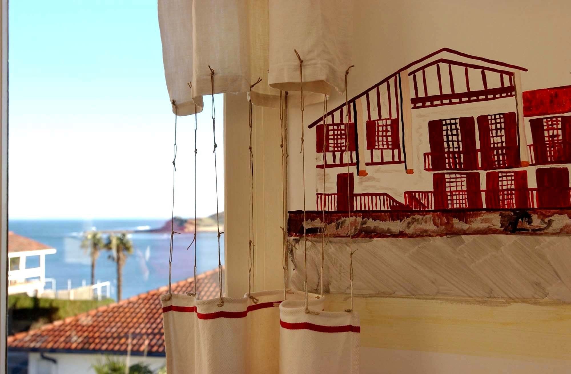 Une pièce offrant une vue sur un paysage côtier, avec une fenêtre donnant sur un bâtiment aux encadrements de fenêtres rouges et doté d’un toit en tuiles.