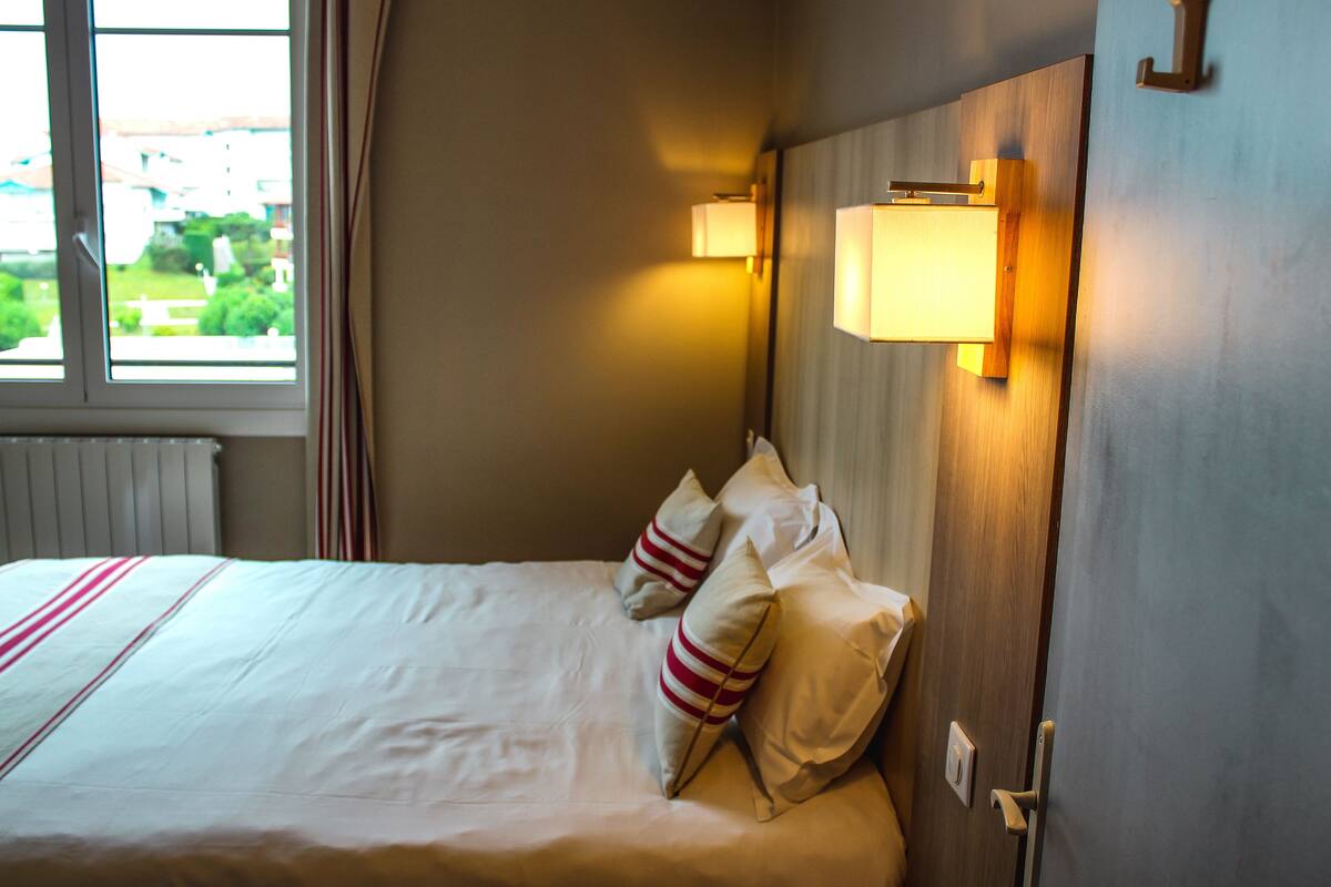 Een hotelkamer met een net opgemaakt bed, twee kussens, een raam met gordijnen en een wandlamp.