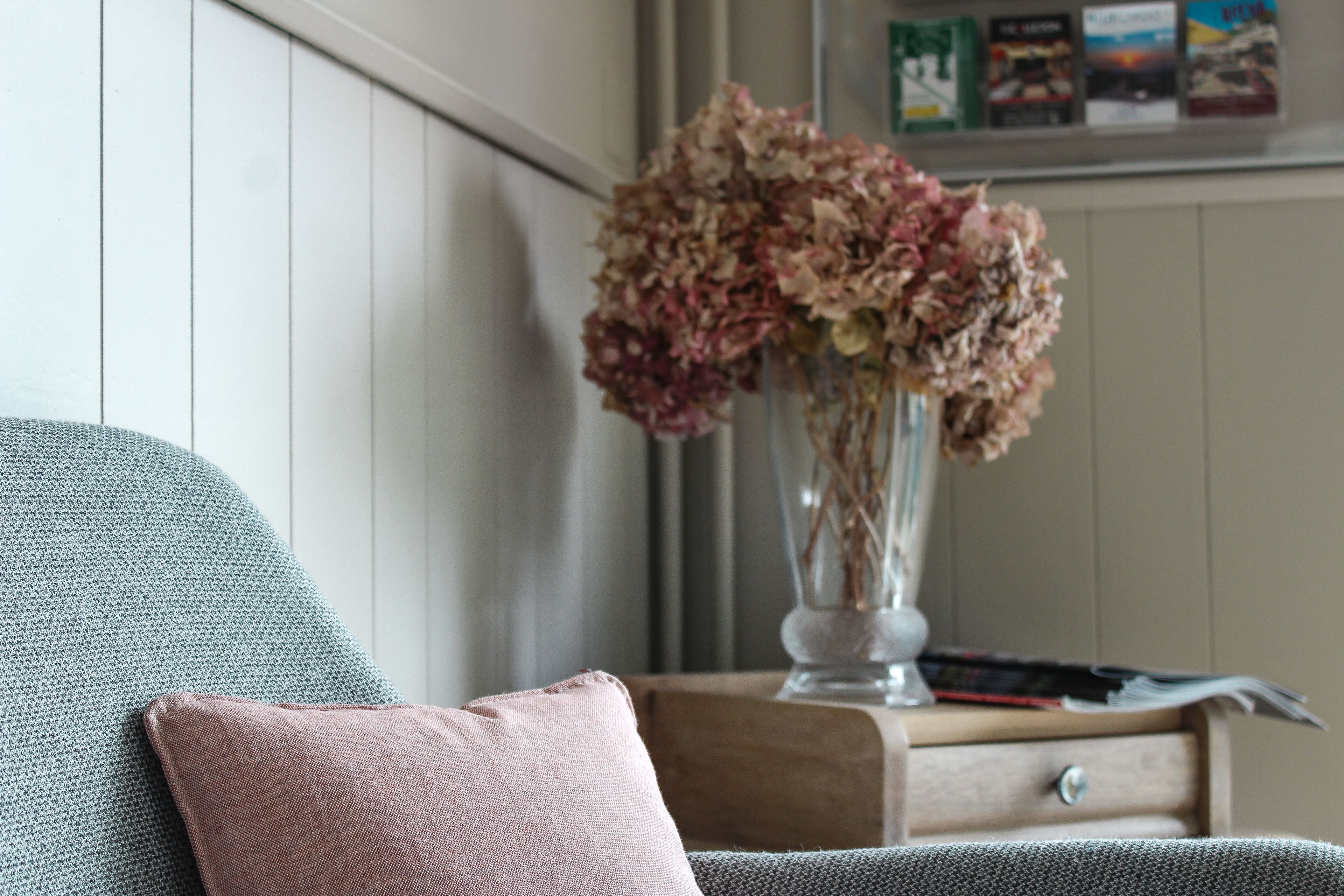 Een kamer met een grijze bank, een houten bijzettafeltje met een vaas gedroogde bloemen, en een prikbord aan de muur.