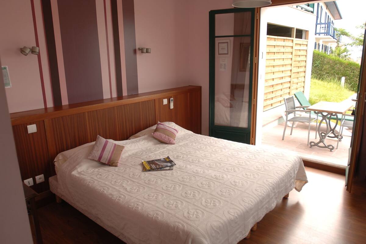 Een slaapkamer met een houten hoofdbord, een bed met een beddekleed met dessin, twee kussens en een boek op het bed. Er is een schuifdeur die leidt naar een buitenruimte met een tafel en stoelen.