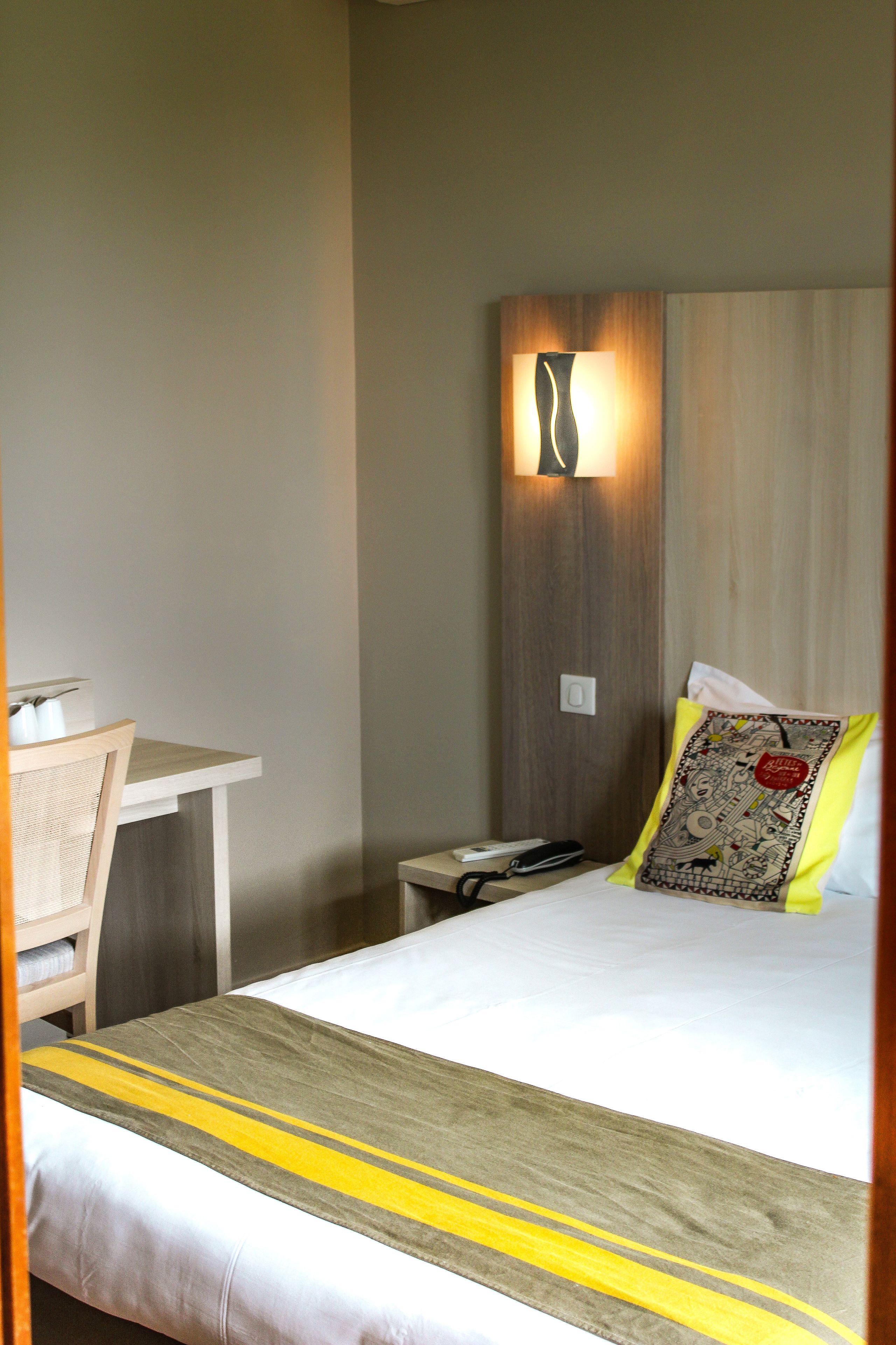 A hotel room with a bed, a bedside table, a chair, and a wall-mounted lamp.