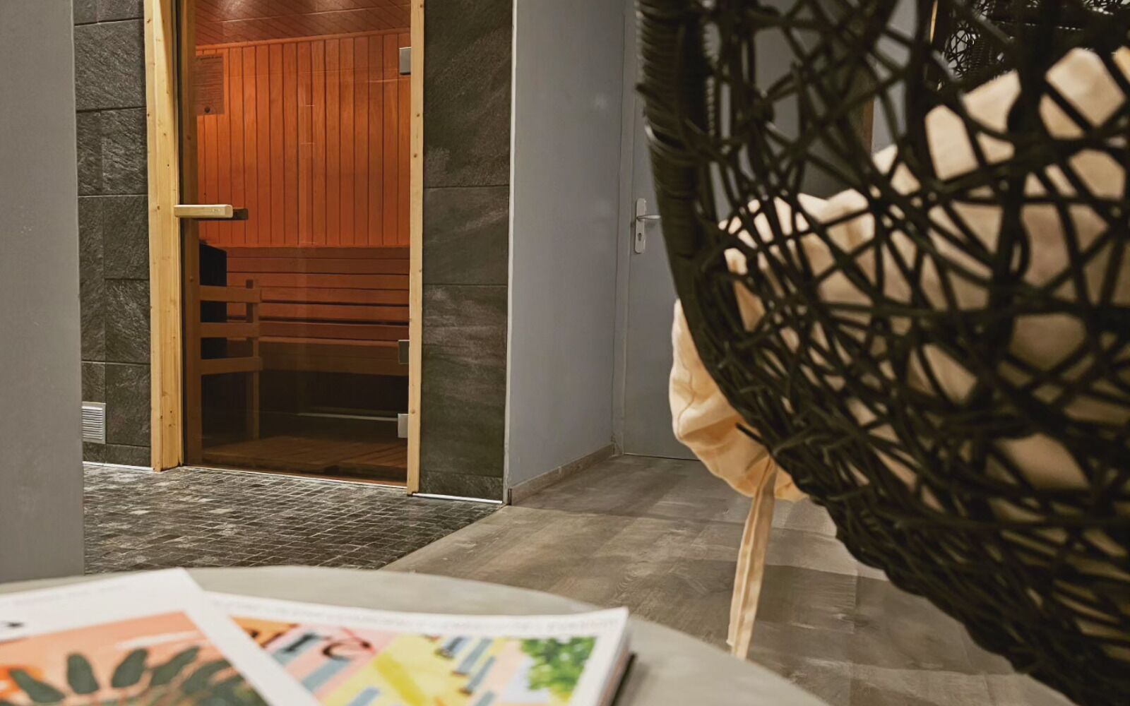 A room with a wooden door leading to a sauna area, a grey wall, and a woven chair.