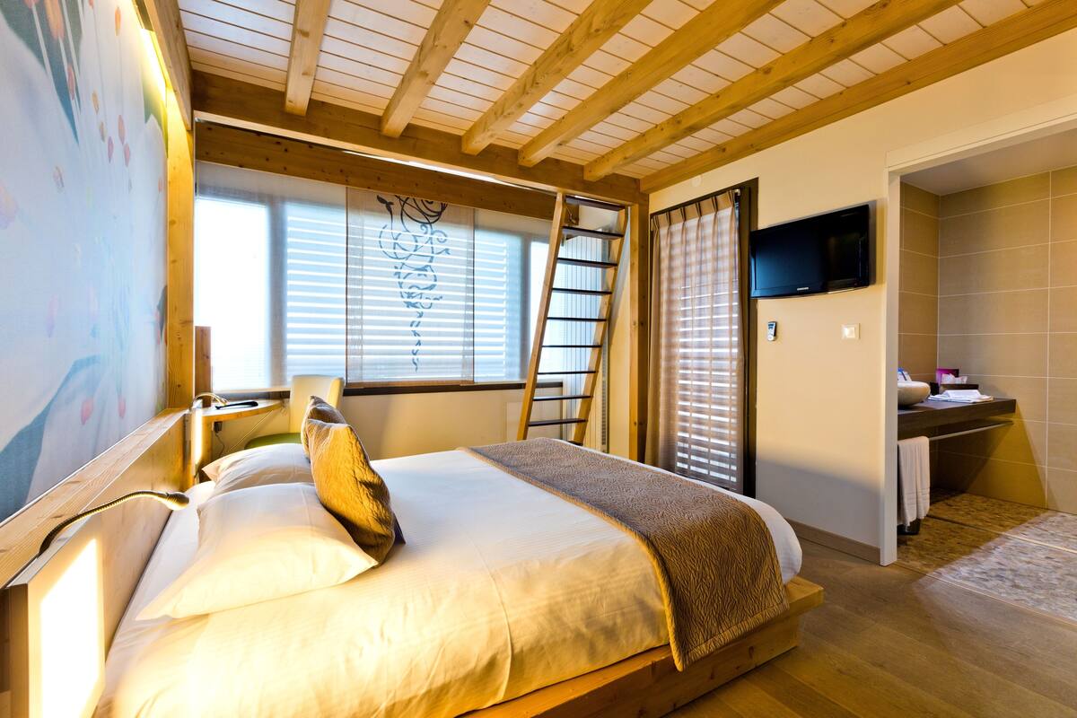 Une chambre à coucher comprenant un lit, un téléviseur fixé au mur, une fenêtre avec des stores, un plafond en bois et une salle de bain avec une baignoire.