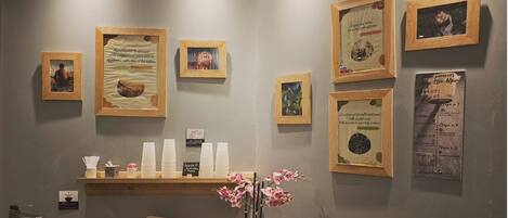Un área de recepción con obras de arte enmarcadas, un estante de madera con tazas blancas, un lavabo de madera y un jarrón con flores rosadas.