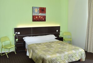 Une chambre d’hôtel avec un lit, deux tables de chevet, une lampe de chevet, un fauteuil vert, un tableau au mur et une fenêtre avec des rideaux.