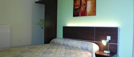 A bedroom with a bed, a nightstand, a lamp, a radiator, and a painting on the wall.