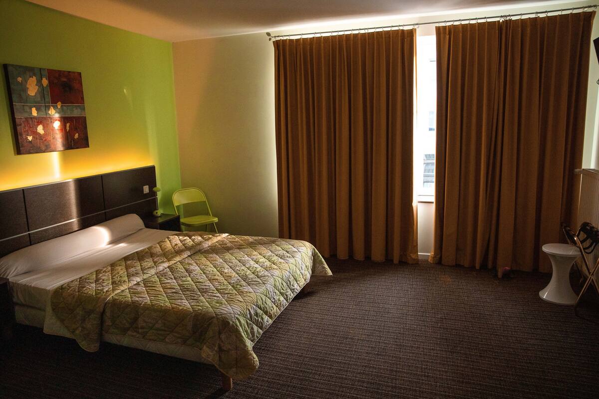 Hotellihuone, jossa on sänky, tuoli, wc ja ikkuna, jossa on verhot.