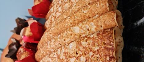 Close-up de uma crepe dobrada com superfície dourada, acompanhada de pétalas de rosa vermelhas e raspas de chocolate.