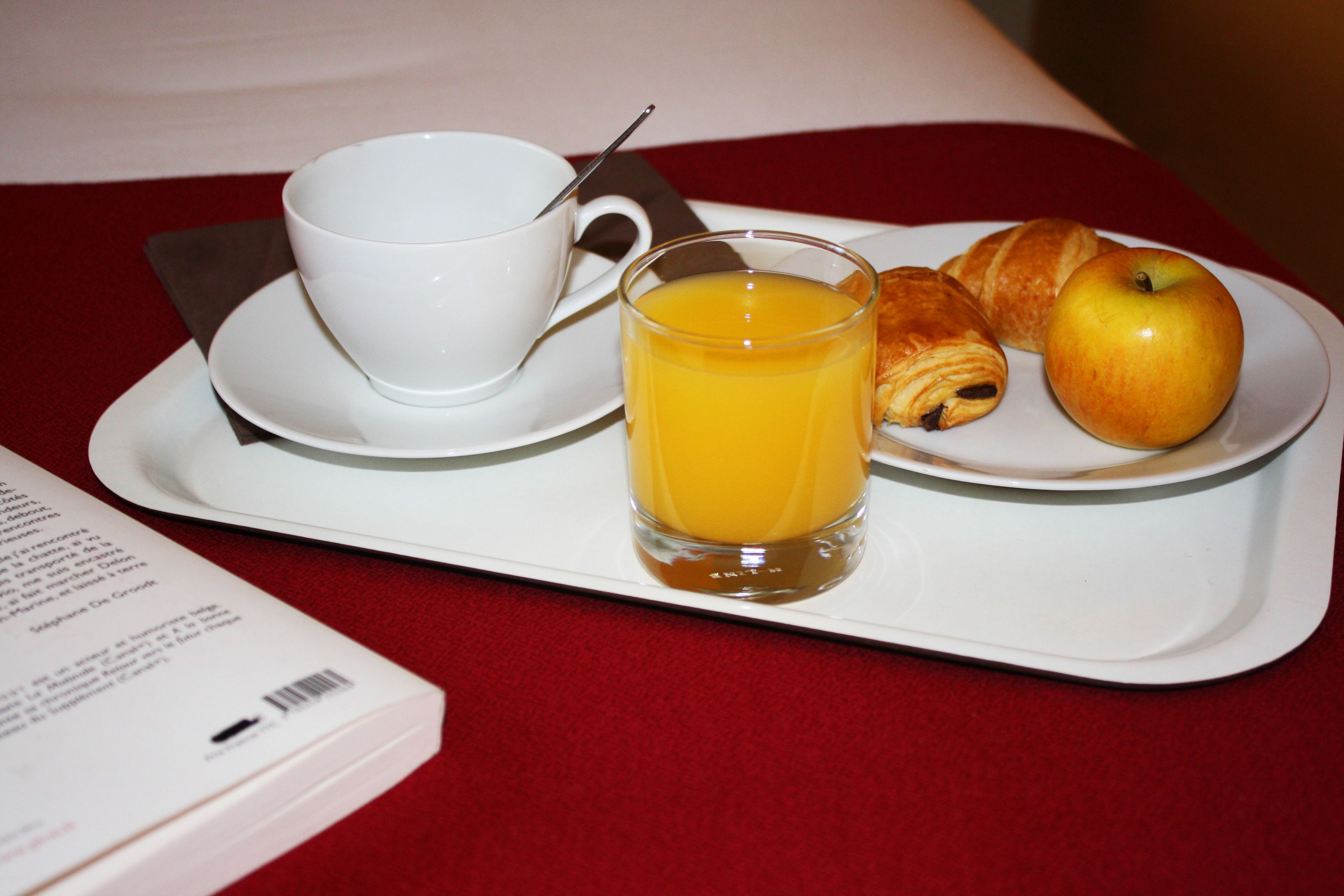 Una bandeja con un croissant, una manzana, un vaso de jugo de naranja y una taza con una cuchara sobre un mantel rojo.