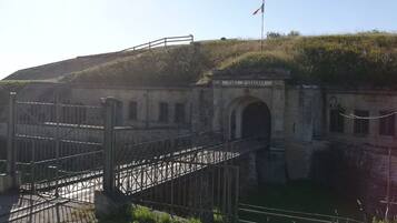 L’entrée d’un fort historique, avec une porte en métal, un drapeau et une colline verdoyante en arrière-plan.