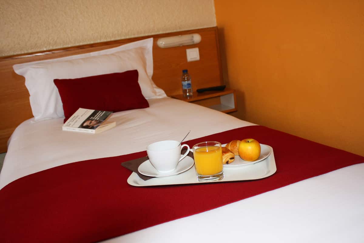 Una cama bien hecha con una manta roja, una bandeja con una taza de café, un vaso de zumo de naranja y un croissant.