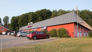 Hotel de fachada roja y tejado gris, estacionamiento con varios coches y árboles al fondo.