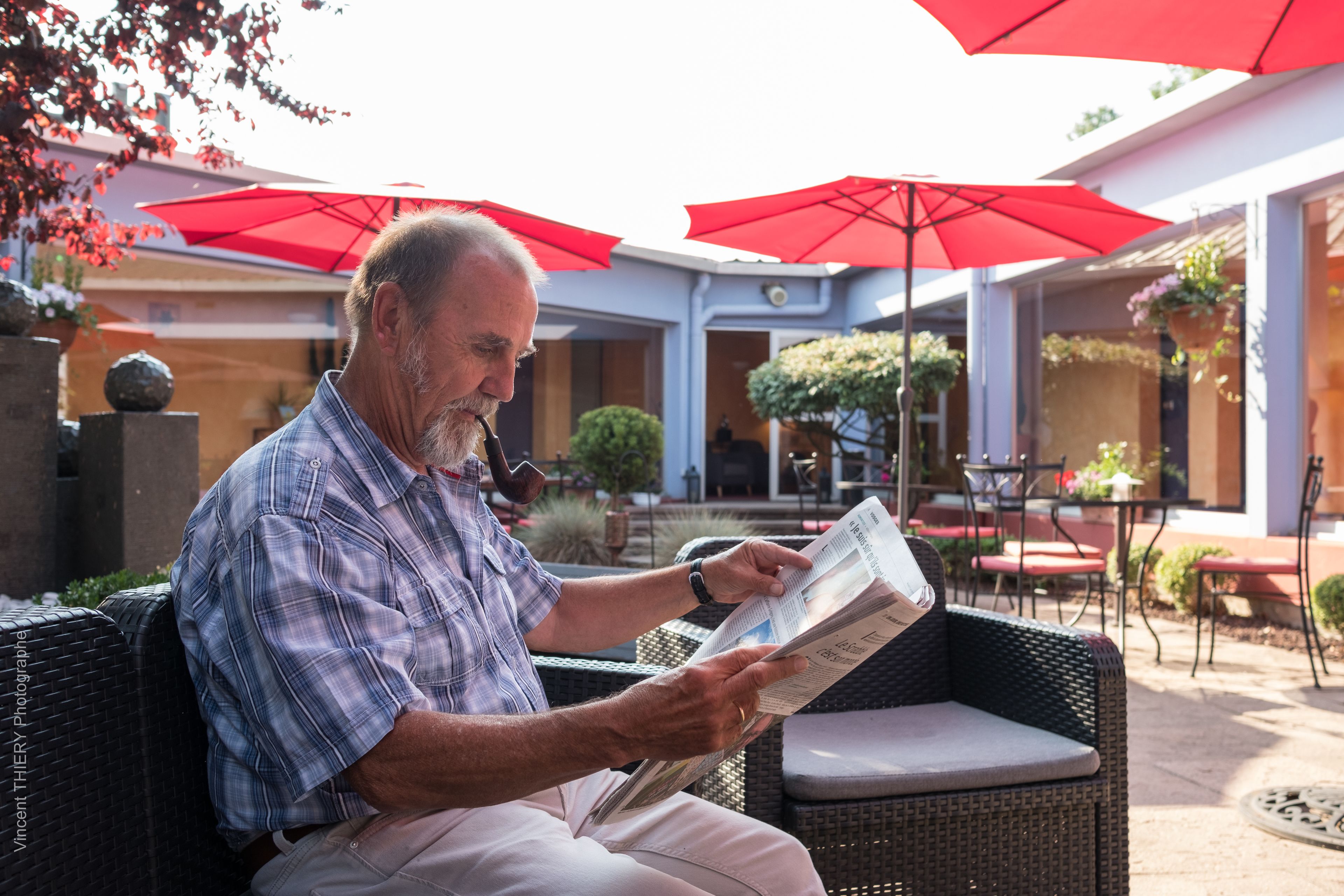 Un homme est assis dans un fauteuil en osier, sous un parasol rouge, et lit un journal.