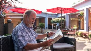 Un homme est assis dans un fauteuil en osier, sous un parasol rouge, et lit un journal.