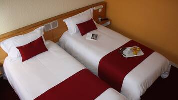 Dos camas individuales con ropa de cama blanca y roja, una mesita de noche con control remoto, y una bandeja con una bebida y un plato de comida.
