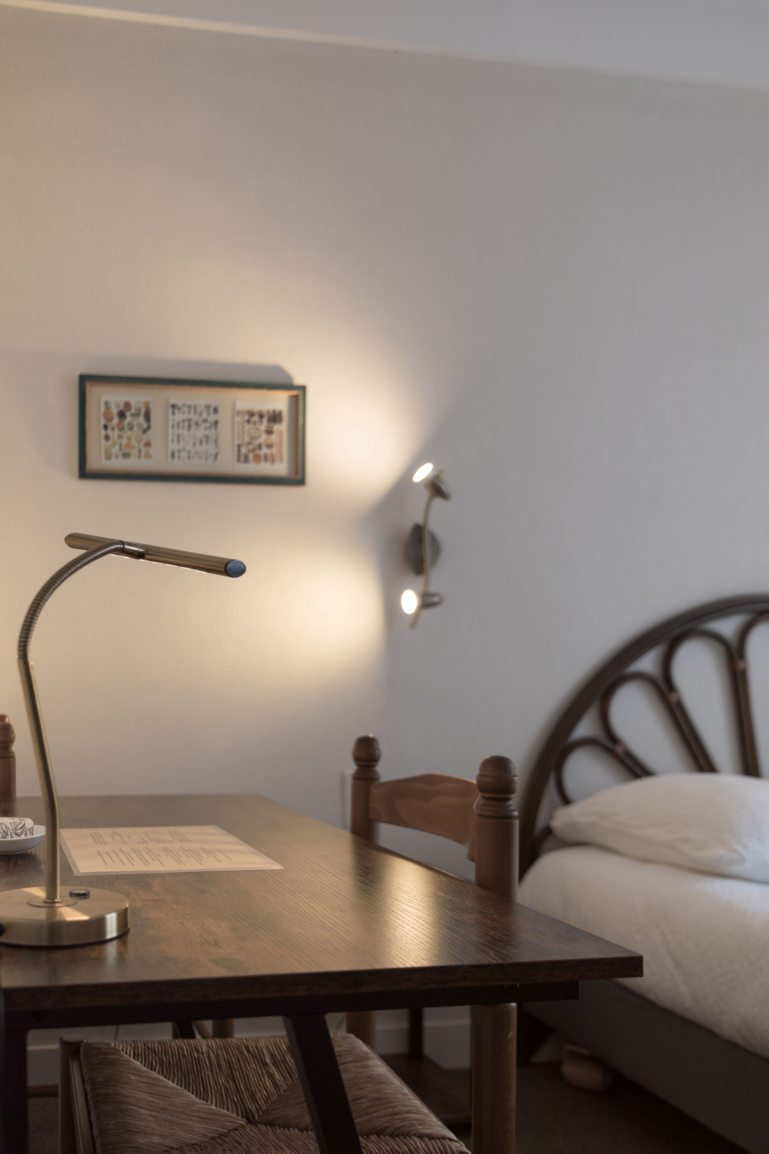 Een kamer met een houten tafel, een bureaulamp, een lijst met een afbeelding aan de muur en een bed met hoofdeinde.
