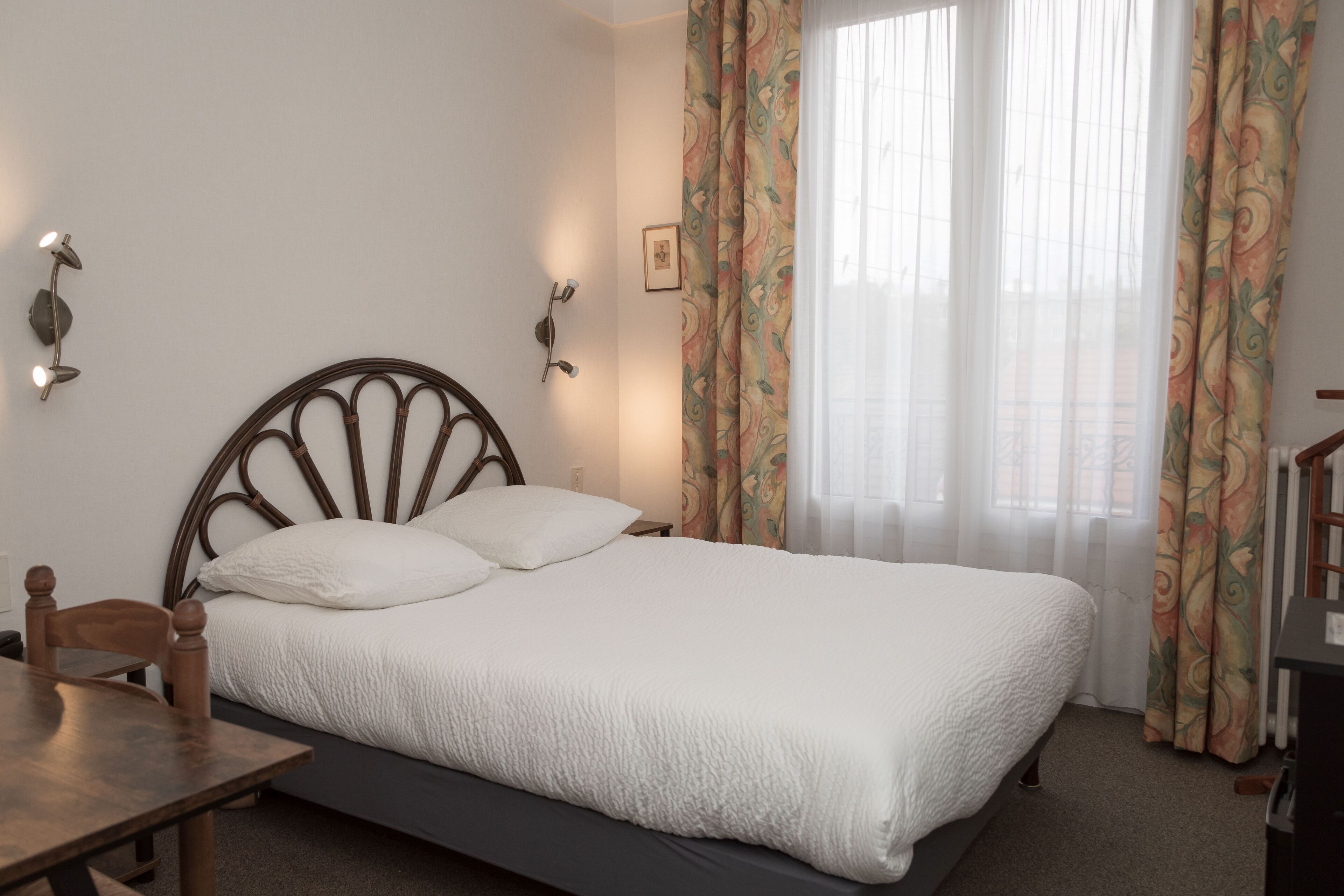 Uma cama de solteiro com cabeceira de madeira, uma mesa de cabeceira e uma cadeira. Há uma janela com cortinas leves e vista para o exterior.