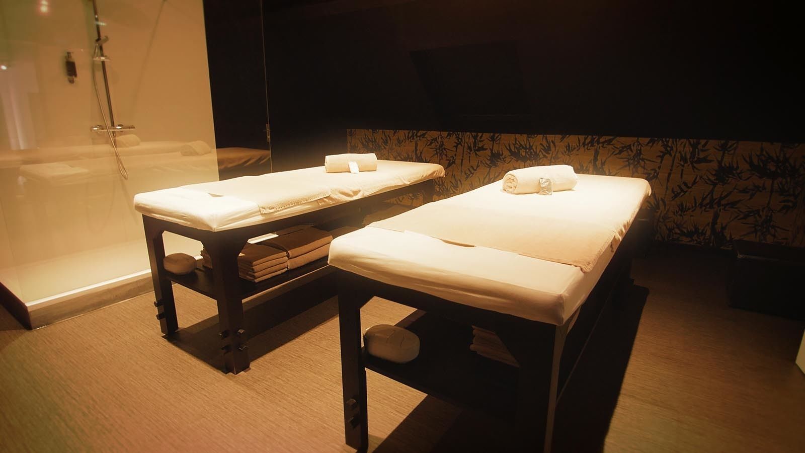 Two massage tables with towels, a glass shower enclosure, and a bamboo-patterned wall.
