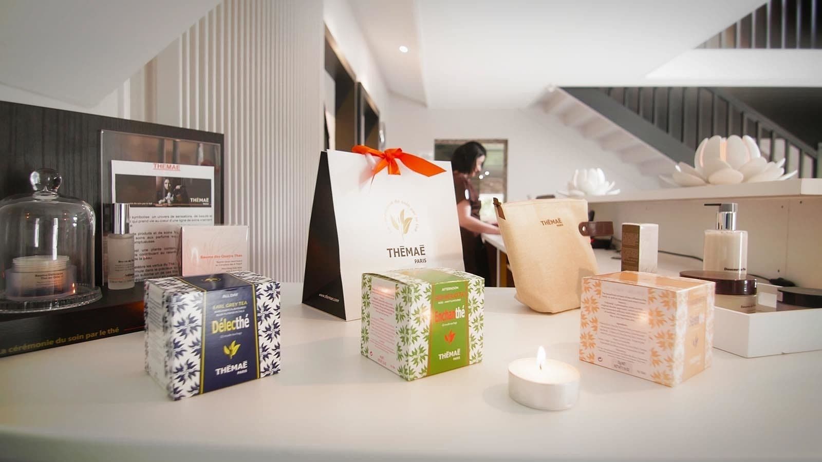 Une présentation des produits Themae Paris, comprenant des boîtes à thé et une bougie, avec un escalier en arrière-plan.