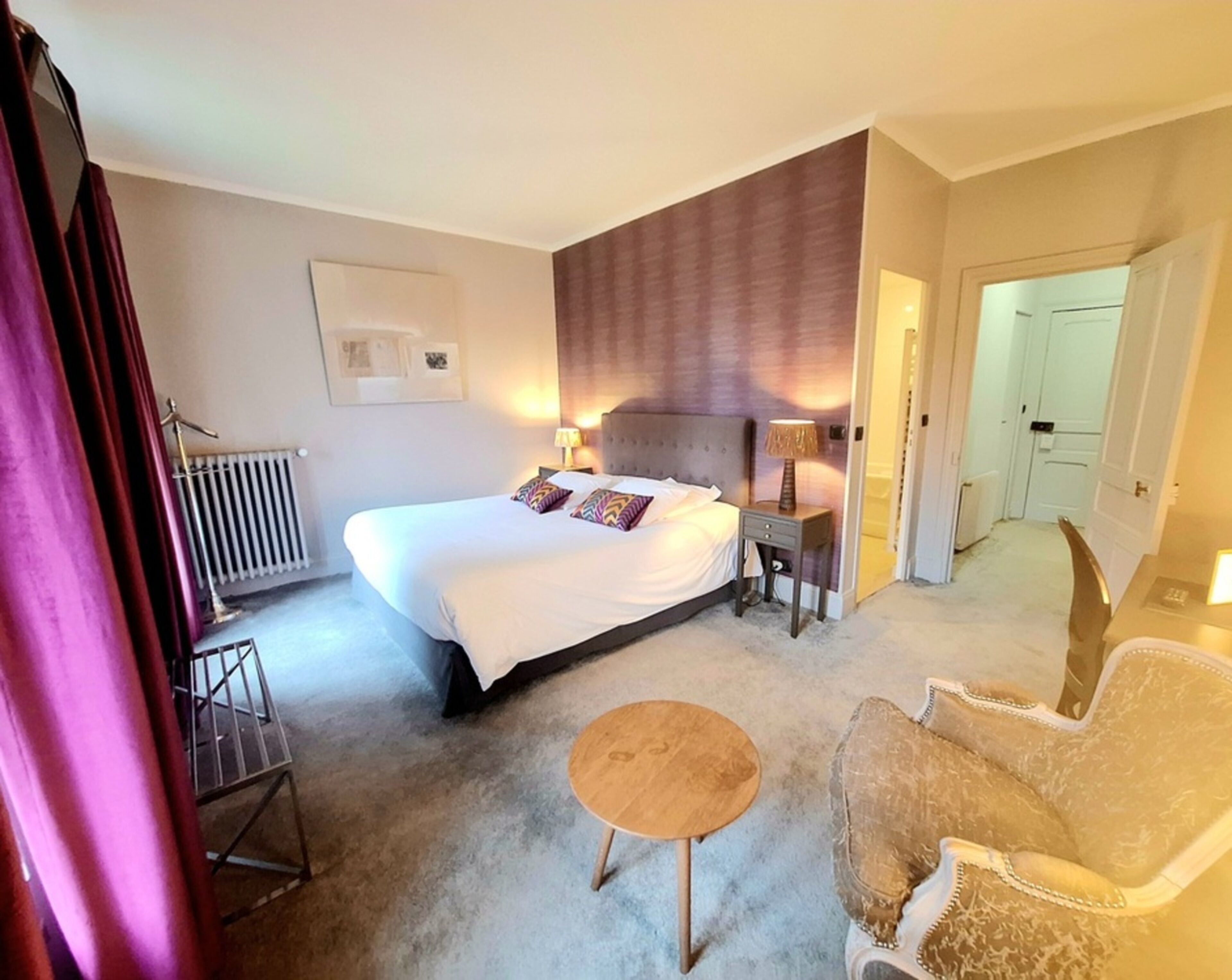 A hotel room with a large bed, bedside tables, a chair, a small round table, and a radiator.