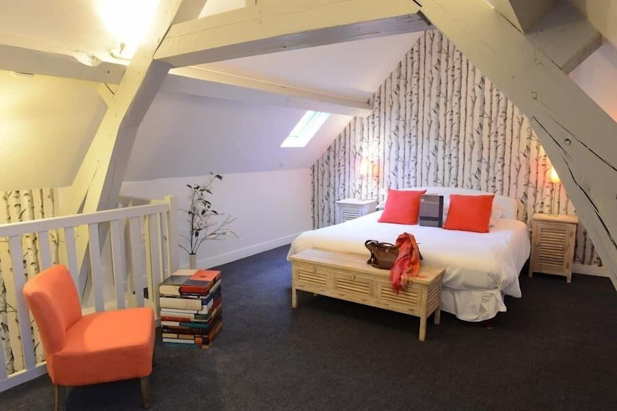 A cozy bedroom with a sloped ceiling, a bed with red and gray pillows, a wooden nightstand, a chair, and a stack of books.