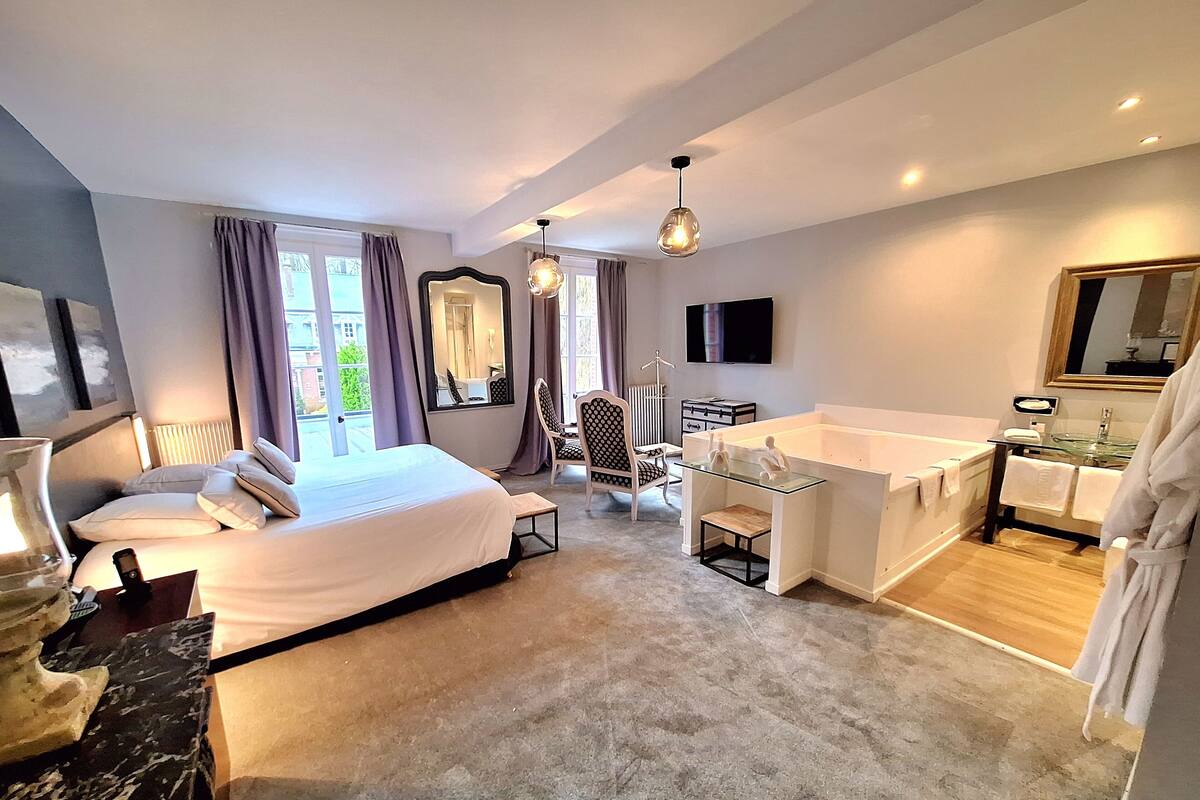 A modern hotel room with a large bed, a glass-enclosed bathtub, a desk with a chair, a large mirror, and a television on the wall.