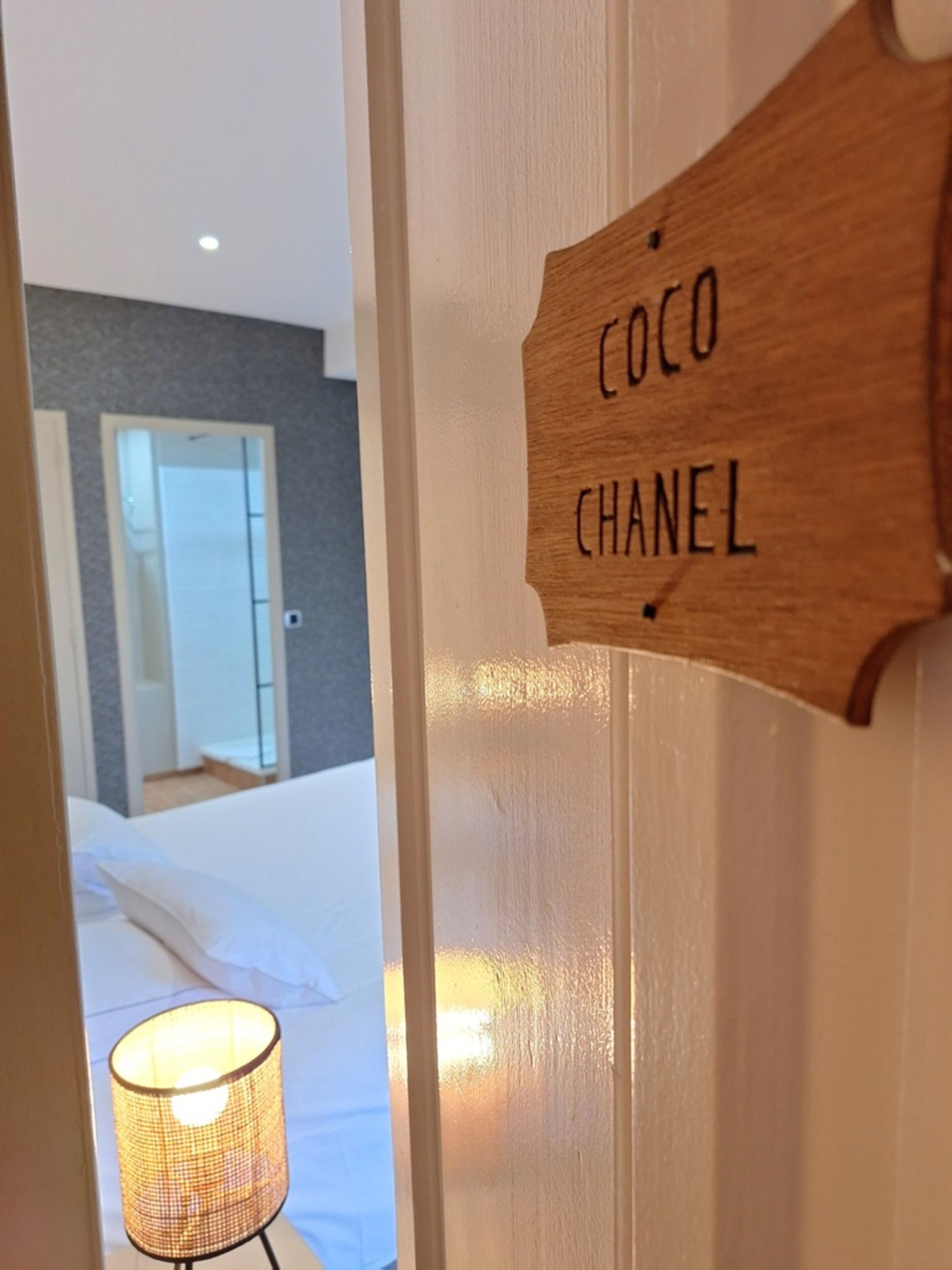 Een houten bord met de tekst 'COCO CHANEL', een bed met wit beddengoed en een lamp met een geweven kap.