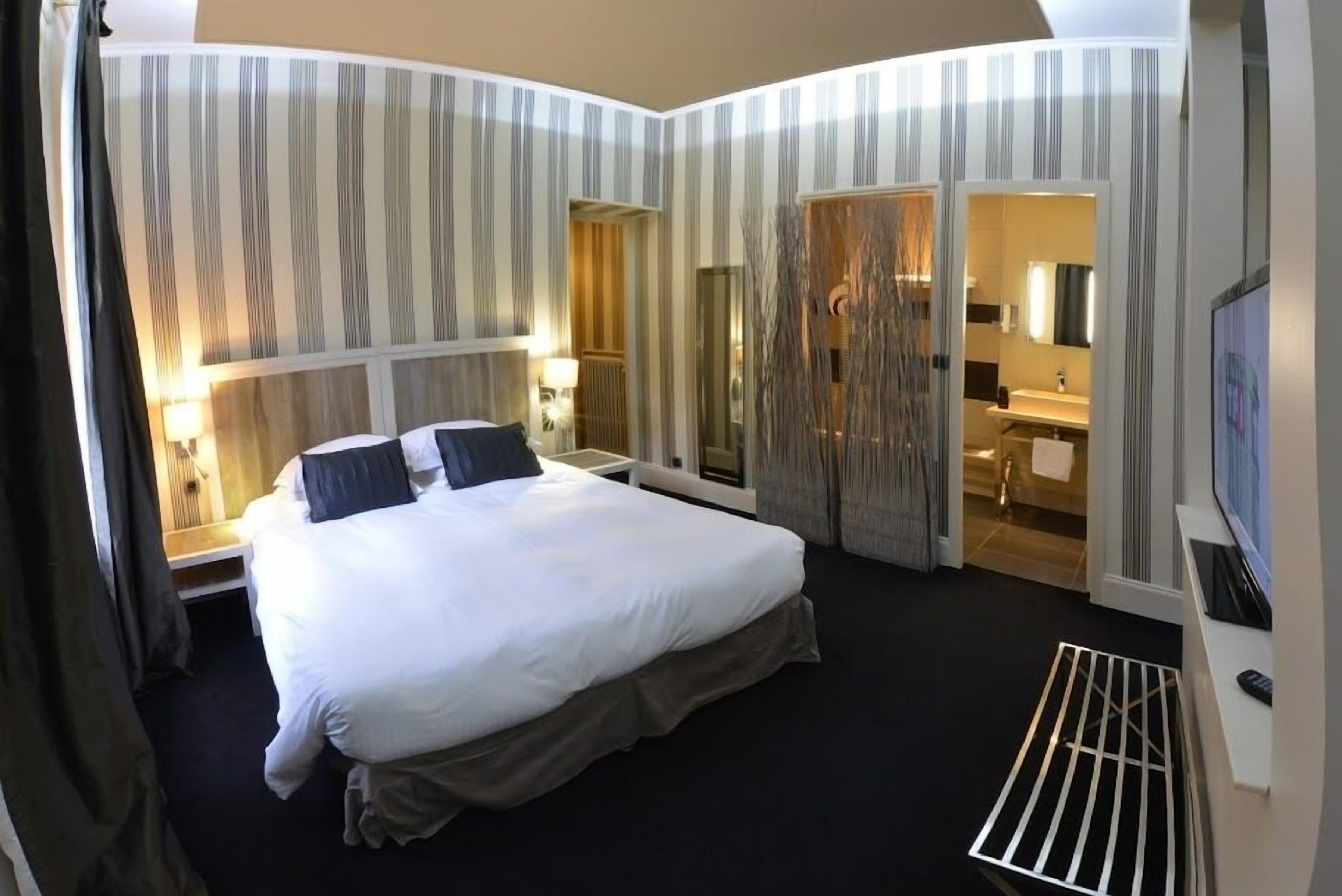 Habitación de hotel con una cama grande, una mesita de noche con lámpara, un espejo, un televisor y una puerta corredera que da al baño.