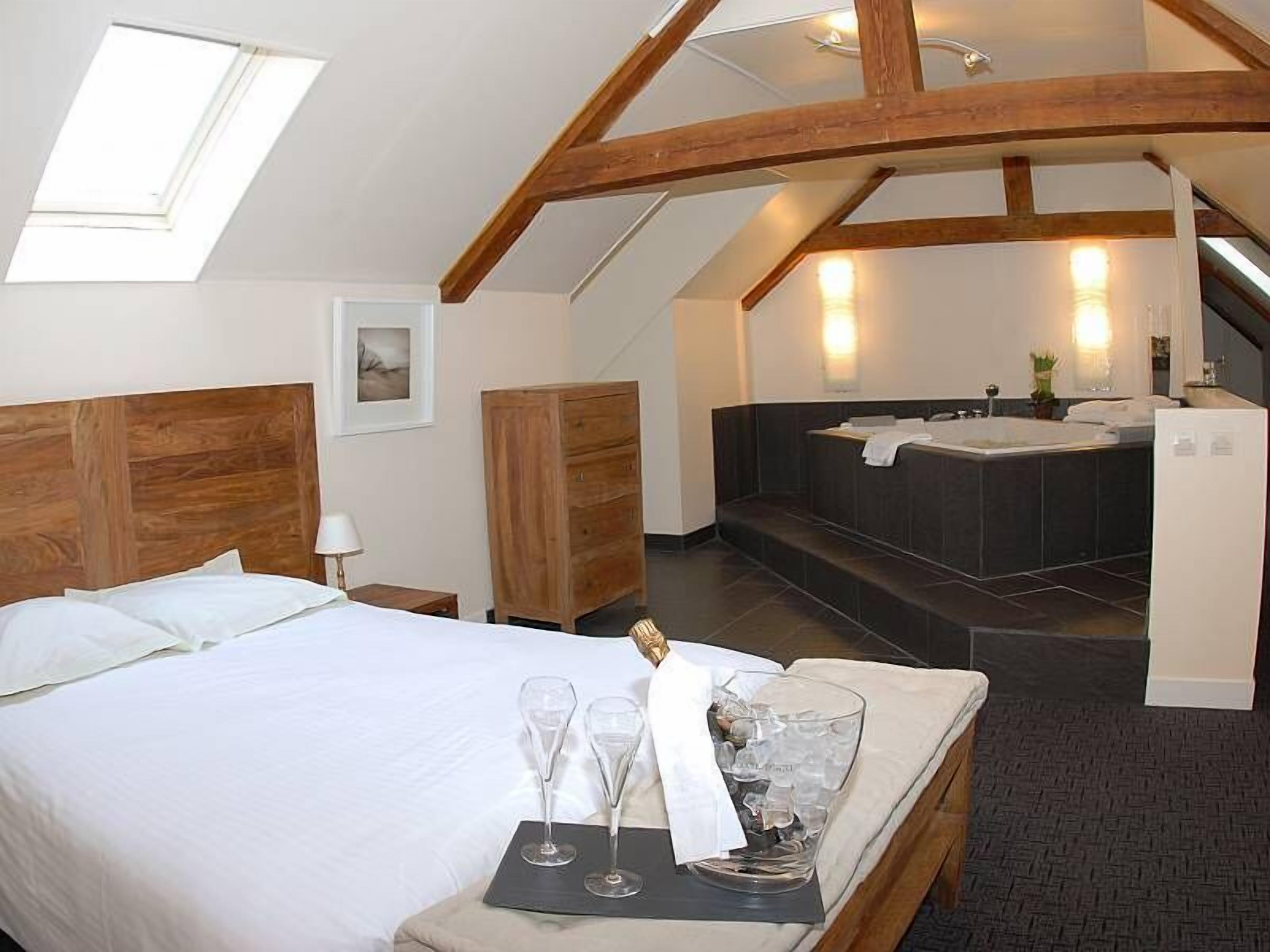 Une chambre d’hôtel comprenant un lit, une tête de lit en bois, une table de chevet, un plateau avec des verres et une bouteille, une salle de bain avec une baignoire et un escalier.
