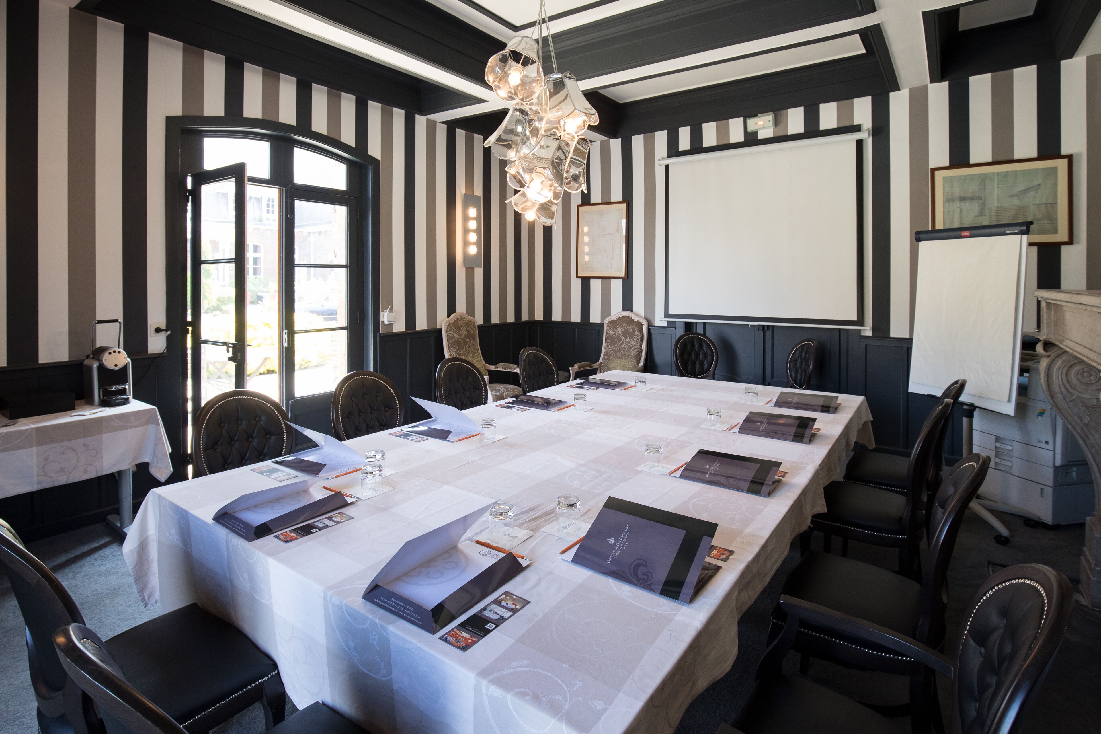 A conference room with a long table, chairs, a projector screen, and a chandelier.