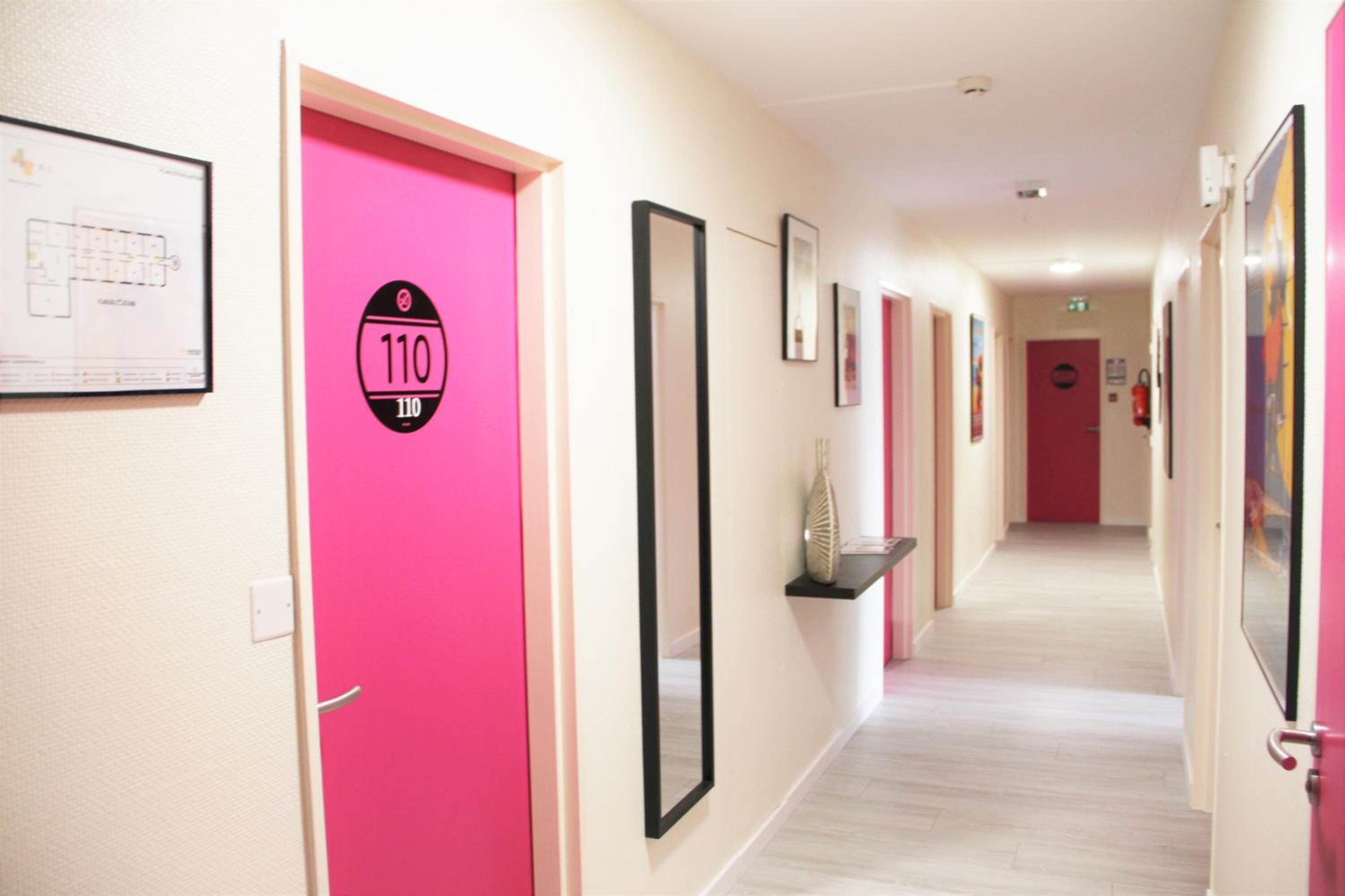 A hotel corridor with pink doors, framed artwork, and a fire extinguisher.