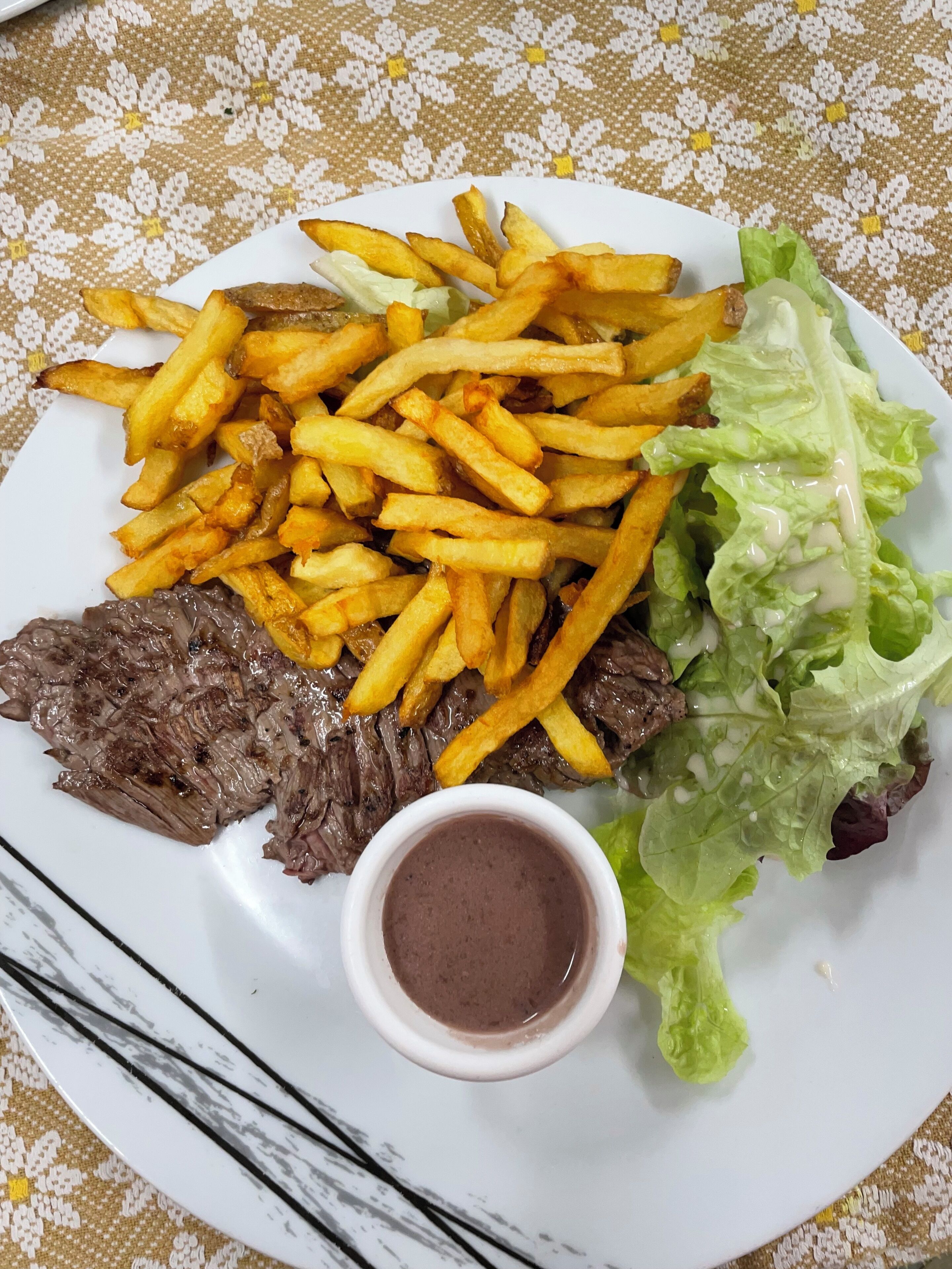 A plate with steak, fries, lettuce, and a small cup of sauce.