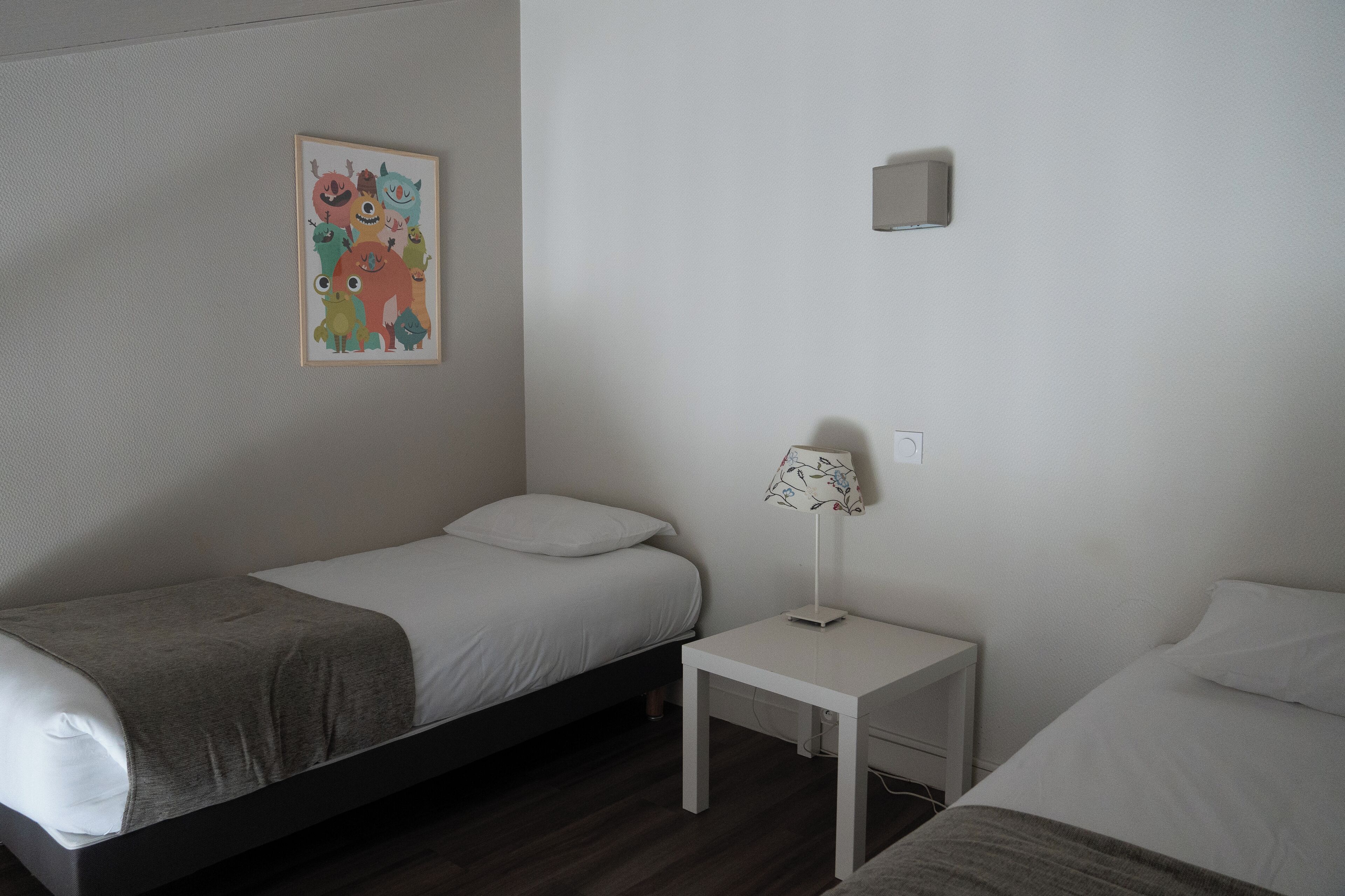 A small, clean room with two single beds, a bedside table with a lamp, and a wall-mounted light fixture.