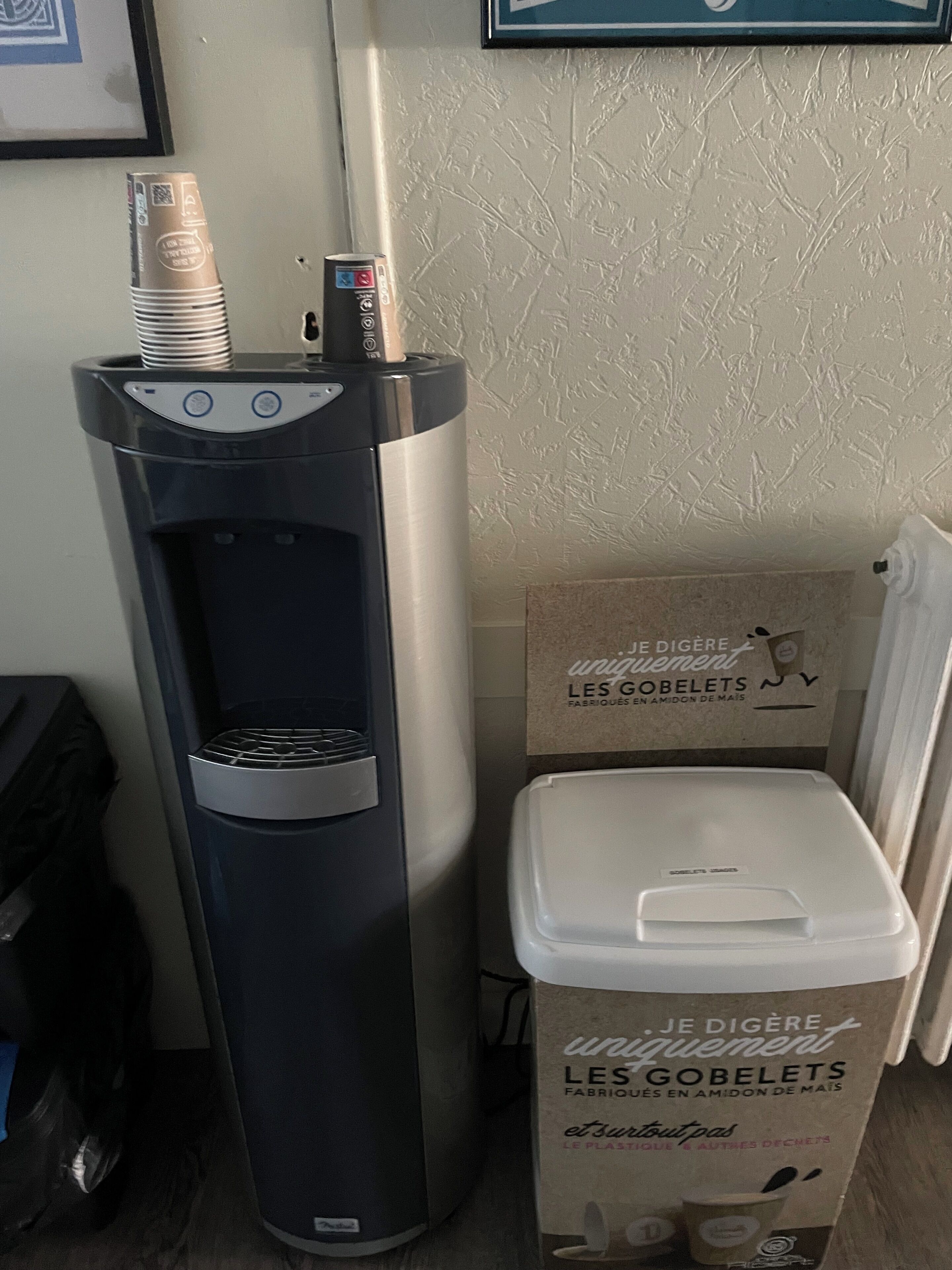 A water dispenser with a coffee machine, a trash bin, and a cardboard box labeled "JE DIGÈRE uniguement LES GOBELETS FABRIÉS EN AMIDON DE MAIS".