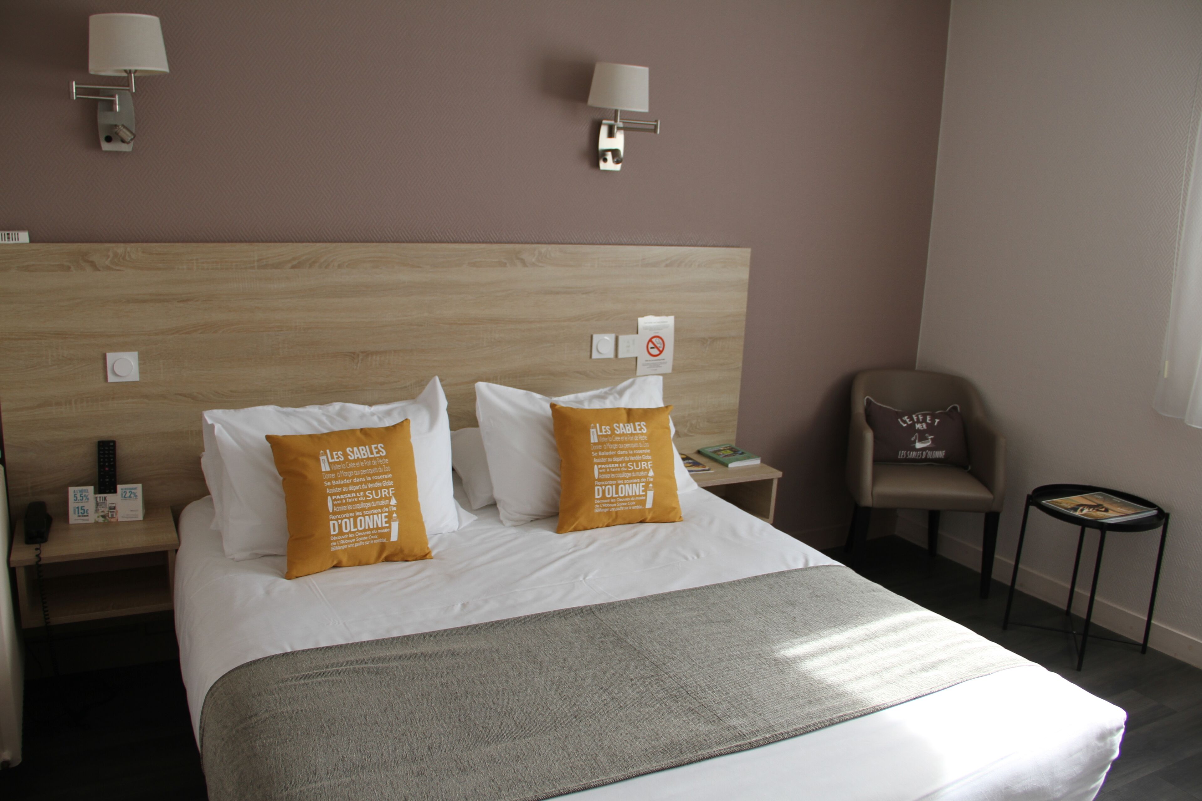 Habitación de hotel con cabecera de madera, dos almohadas blancas, una manta gris, una silla, una mesita de noche con un teléfono y una lámpara.