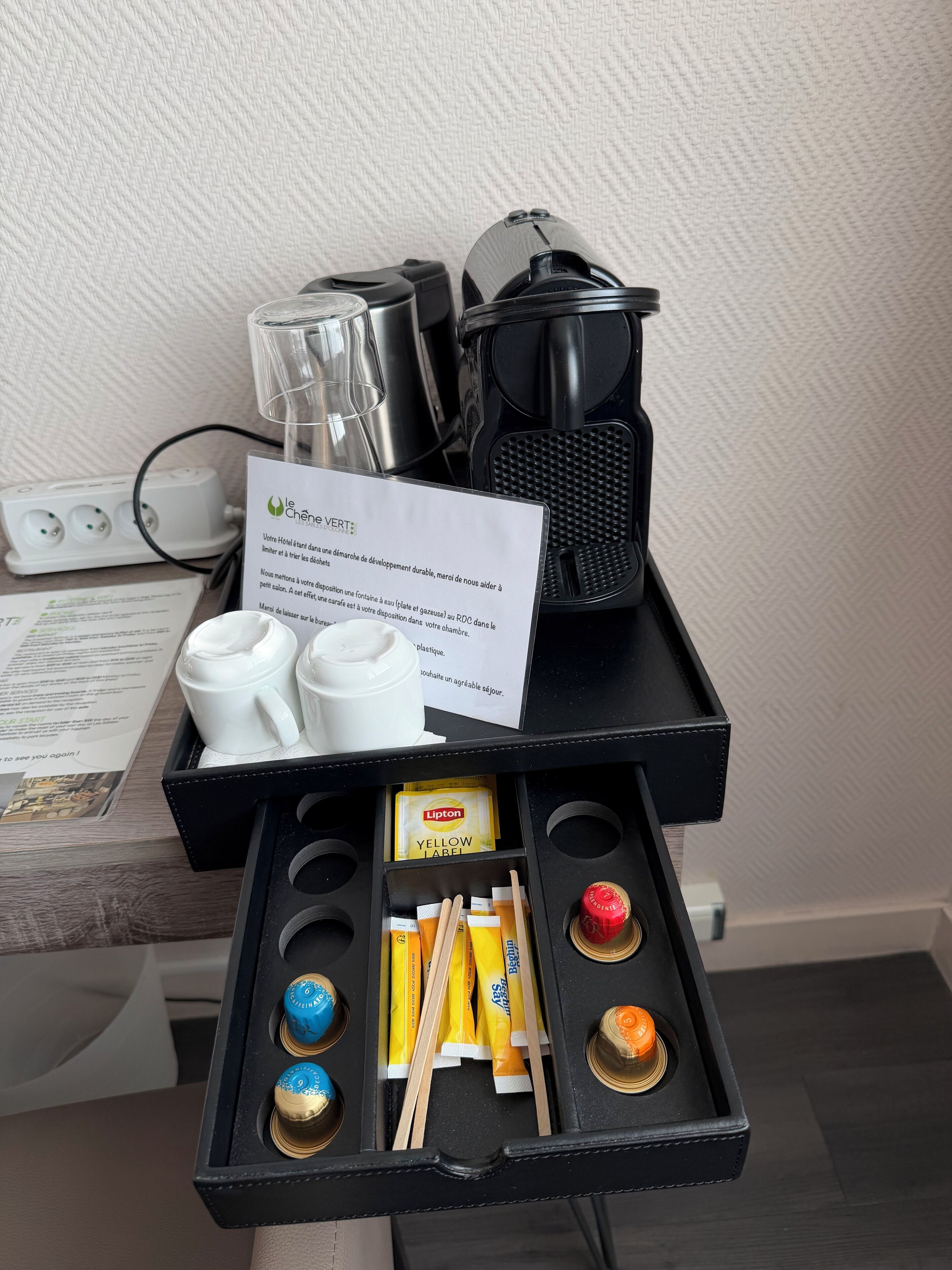 Un mini-bar d’hôtel comprenant une machine à café, un service à thé, des sachets de thé Lipton et un petit plateau avec des condiments.