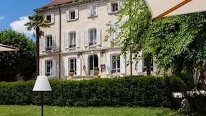 A patio area with outdoor furniture, a hedge, and a building with multiple windows and shutters in the background.
