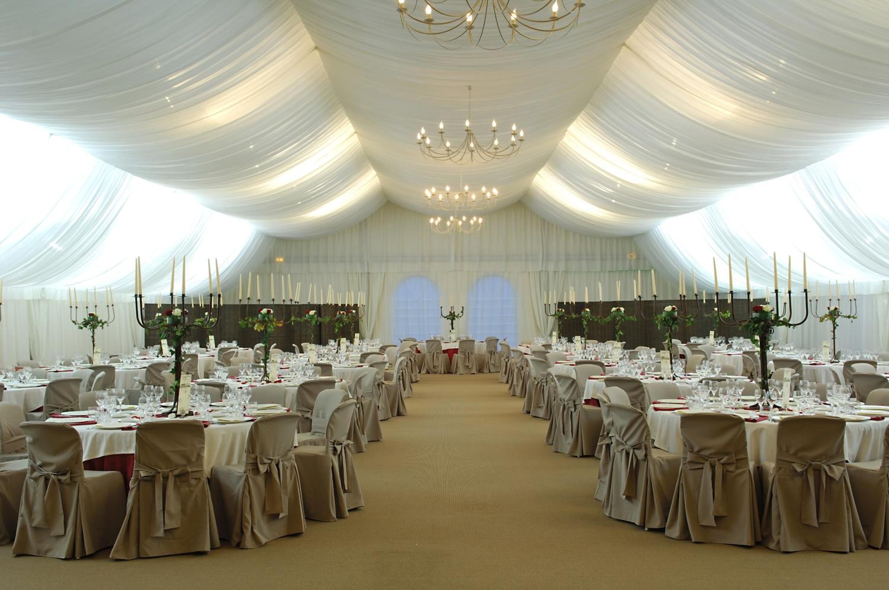 Una sala da banchetti con tavoli rotondi apparecchiati per un evento formale, decorata con drappeggi bianchi e lampadari.