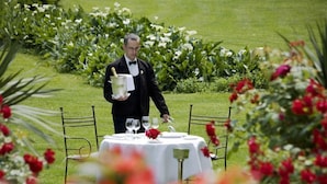 A waiter in a black suit holding a champagne bottle, standing by a table set with white tablecloths and red flowers, surrounded by greenery and flowers.