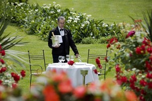 Un camarero con traje negro sostiene una botella de champán, de pie junto a una mesa con mantel blanco y flores rojas, rodeado de follaje y flores.