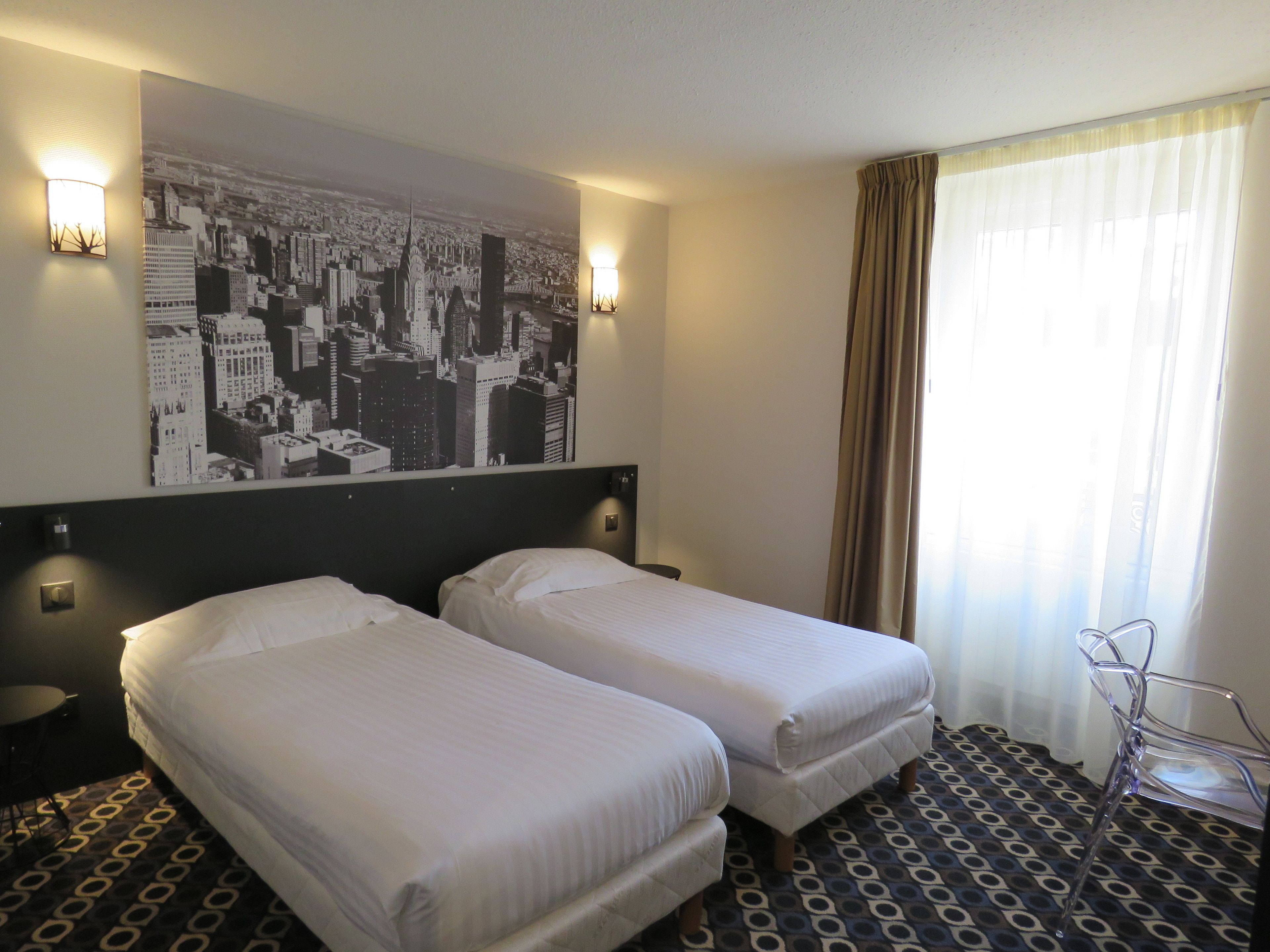 A hotel room with two single beds, a cityscape poster on the wall, and a patterned floor.